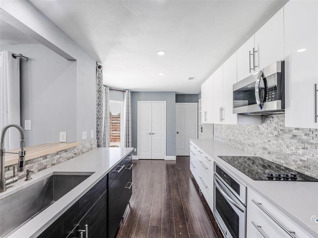 Modern kitchen featuring wood-finish flooring, white flat-panel upper cabinetry, dark flat-panel lower cabinetry, light-toned countertops, and a full-height tile backsplash.  The pantry is at the left double doors and the laundry room is beyond the door to the right. Note teh large sink!