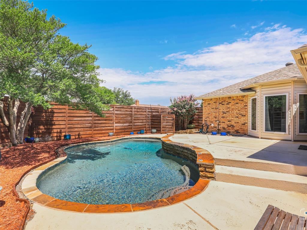 Curved swimming pool with stone coping, surrounded by a concrete patio and flower beds.