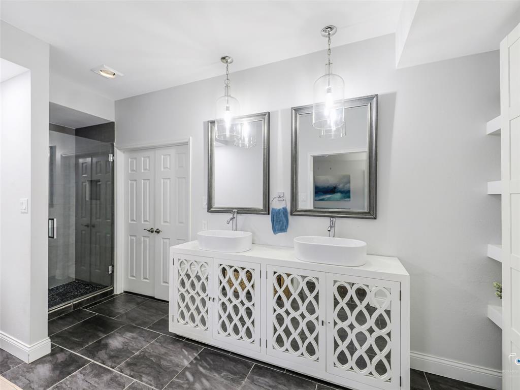 Contemporary bathroom featuring a double vanity with vessel sinks and decorative cabinet fronts, tile floors and a large walk-in shower.  The spacious primary closet is beyond the double doors.