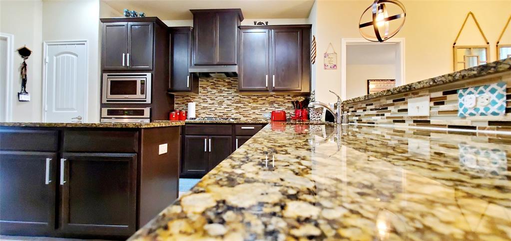 Spacious kitchen featuring expansive stone countertops, dark wood cabinetry, a mosaic tile backsplash, built-in stainless steel appliances, and a kitchen island with additional storage