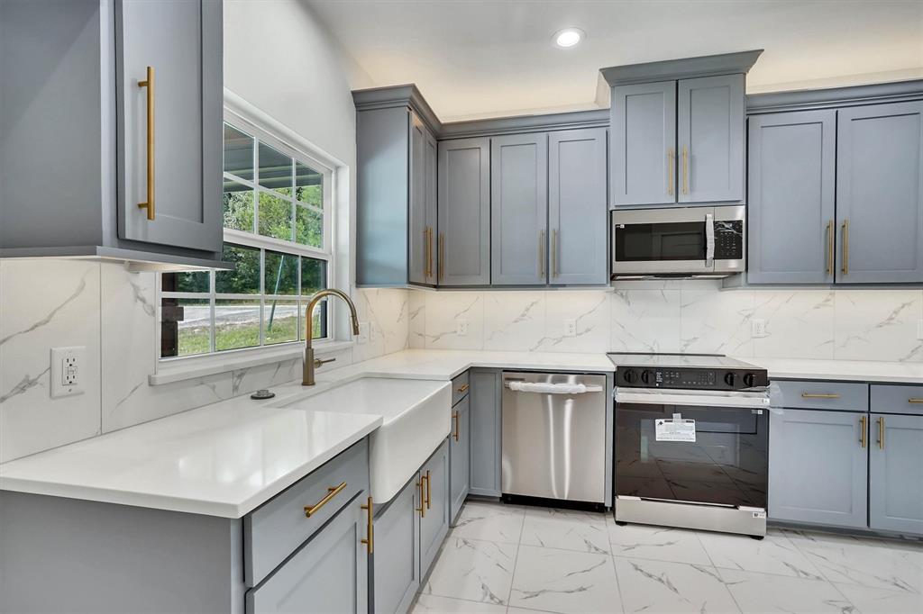 Modern kitchen featuring light gray blue cabinetry, gold-tone hardware, quartz white countertops, a white farmhouse sink, and a stainless steel appliance package