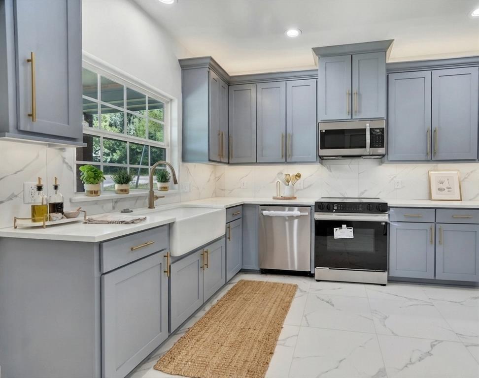 Modern kitchen featuring light gray cabinetry with gold hardware, white countertops, a deep farmhouse sink, stainless steel appliances, and white marble-look tile flooring