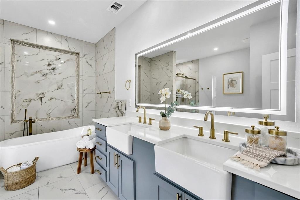 Contemporary bathroom featuring a freestanding tub, dual farmhouse sinks, light gray-blue vanity cabinetry, and a large backlit mirror