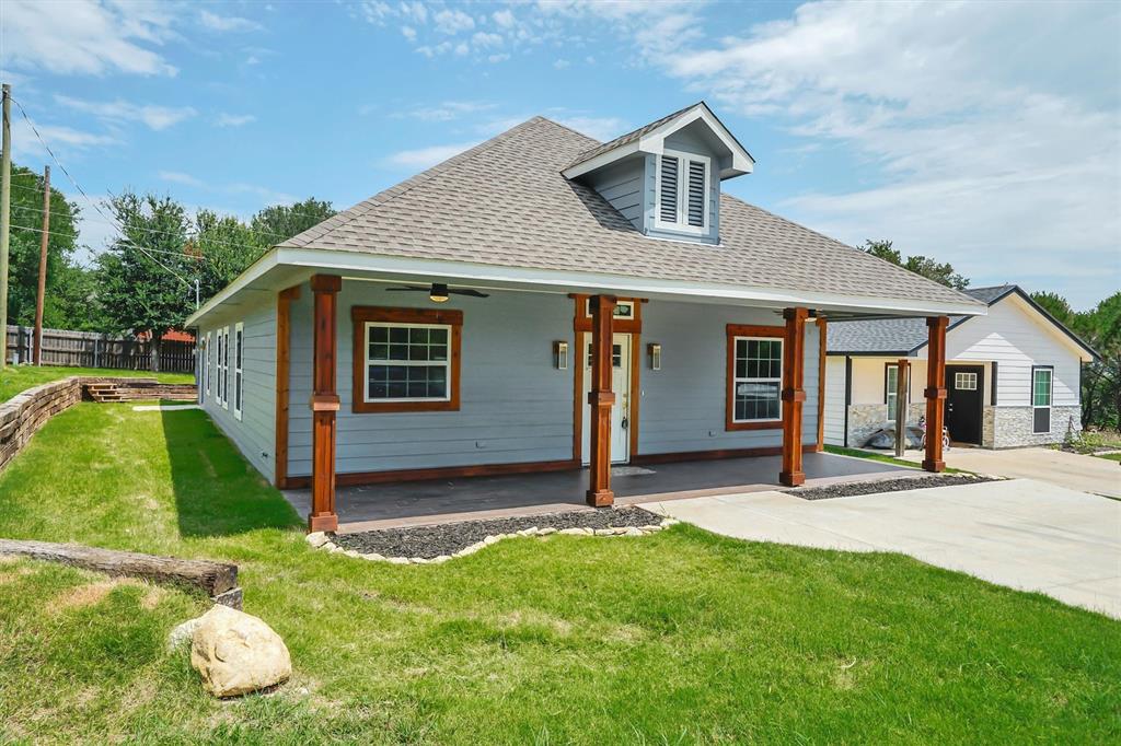Gable roofline with a dormer, covered front porch, exposed wood-finish support columns, concrete driveway, and a tiered landscaped yard