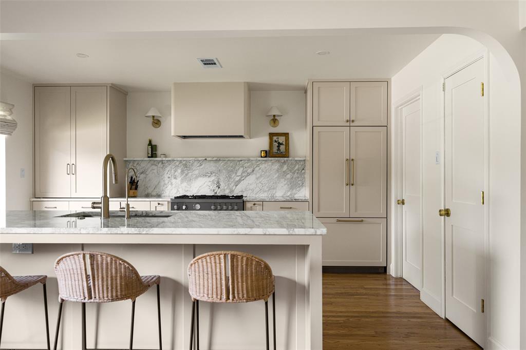 Kitchen featuring light-toned cabinetry, a large center island with a built-in sink, stone countertops and backsplash, a professional-grade range with a custom hood, and integrated refrigeration