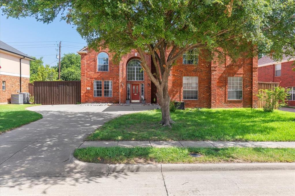 Traditional brick exterior featuring an arched entryway with a red door, multiple large windows, a mature front yard tree, and a concrete driveway