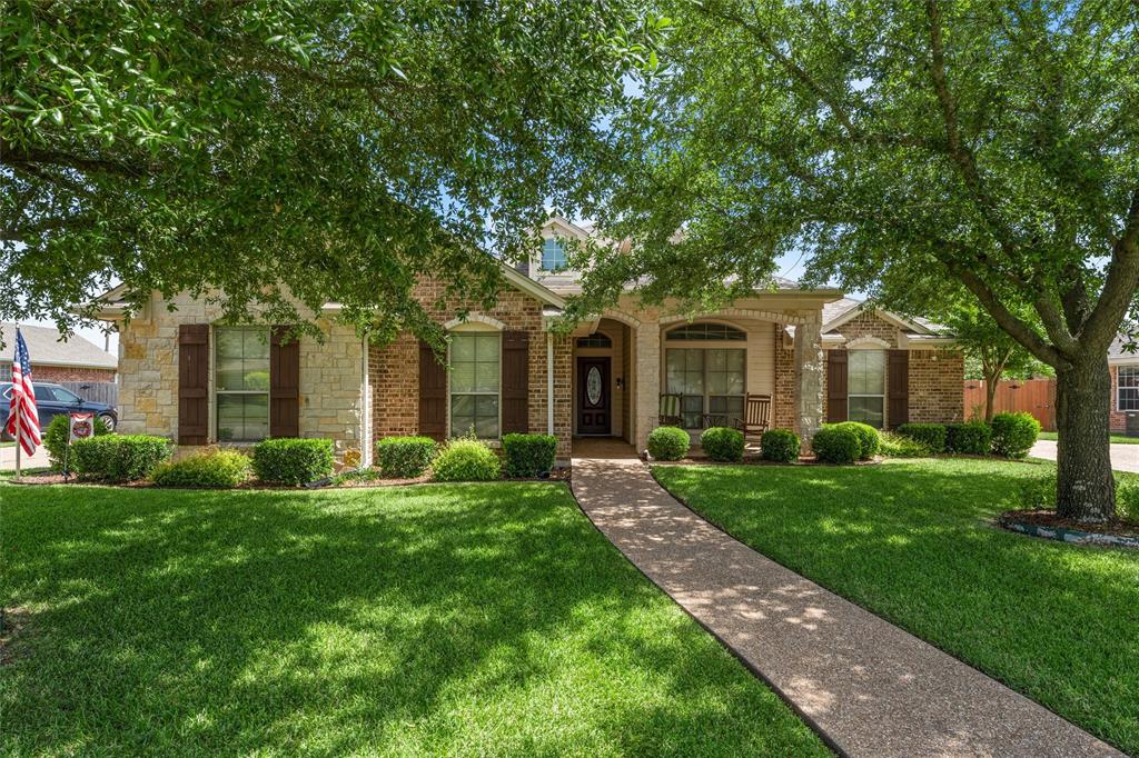Brick and stone facade residence featuring a covered entry porch, dark wood shutters, a well-maintained lawn, and mature trees providing natural shade