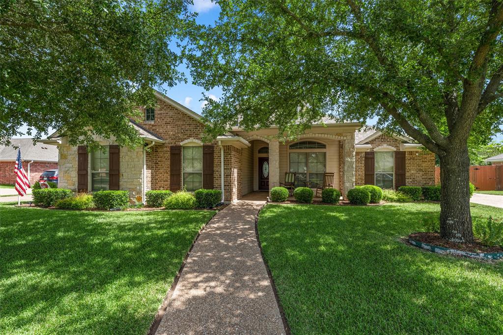 Brick and stone facade residence featuring a covered front porch, mature trees, manicured lawn, and a patterned concrete walkway Brick and stone facade residence featuring a covered front porch, mature trees, manicured lawn, and a patterned concrete walkway