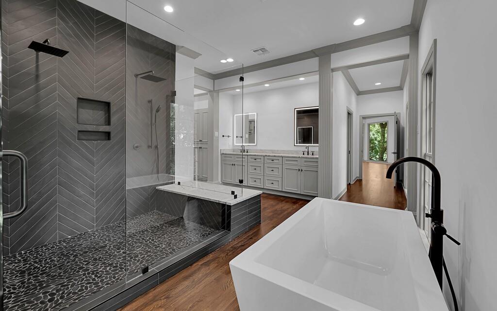 Modern bathroom featuring a freestanding soaking tub with a matte black floor-mounted faucet, a spacious glass-enclosed shower with dark chevron-patterned tile, and a dual vanity with light gray cabinetry and integrated sinks