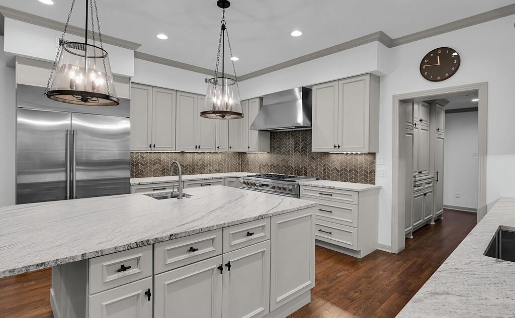Gourmet kitchen featuring a large central island with an undermount sink, stainless steel appliances, light-toned cabinetry with dark hardware, a herringbone pattern backsplash, and wood-finish flooring