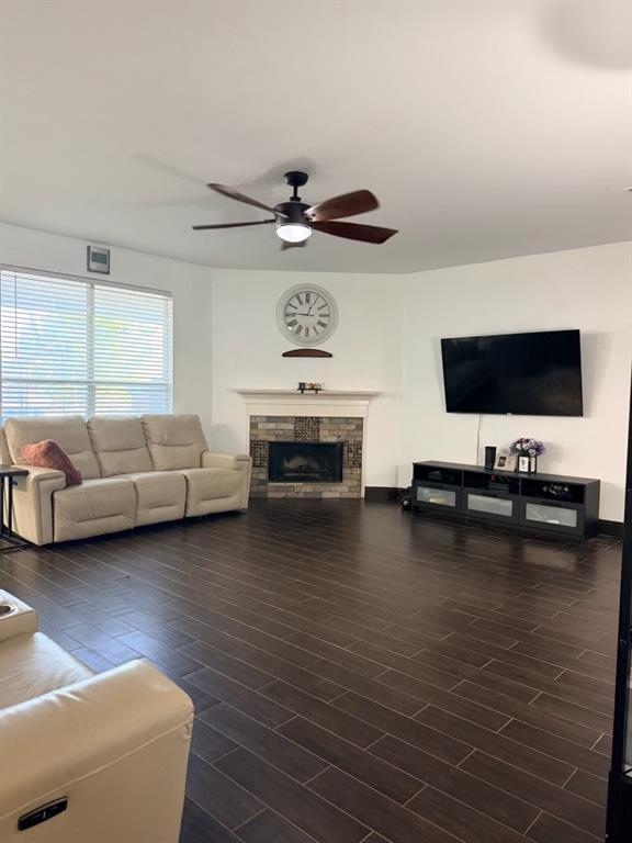 Living room featuring wood-finish flooring, a corner fireplace with stone surround and mantle, and a ceiling fan with integrated lighting