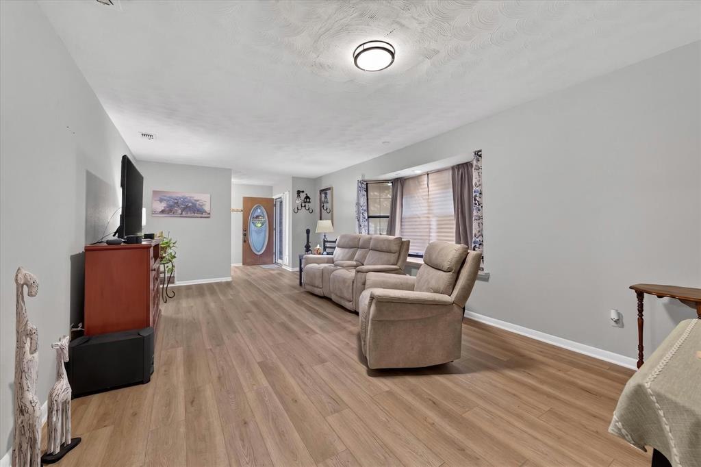 Spacious living area featuring wood-finish flooring, light gray walls, and a recessed bay window with blinds Spacious living area featuring wood-finish flooring, light gray walls, and a recessed bay window with blinds