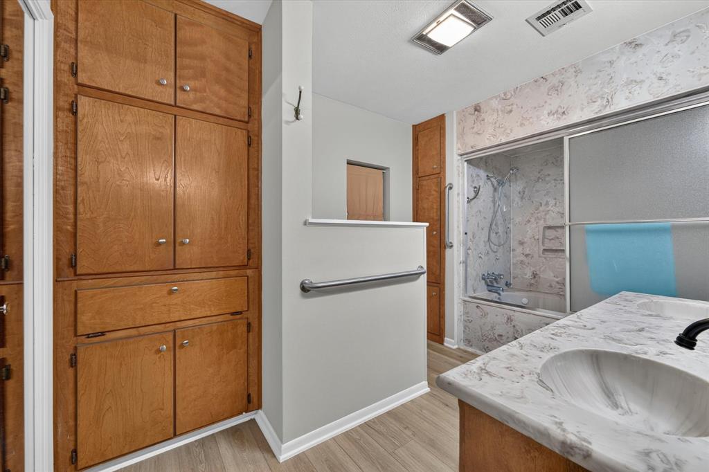 Bathroom featuring a built-in wood-finish storage cabinet, a vanity with an integrated sink and patterned countertop, and a shower-over-tub combination with a sliding glass enclosure