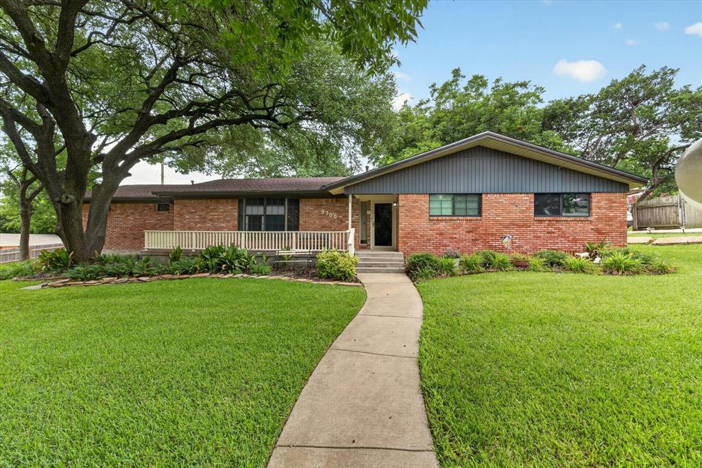 Brick exterior home featuring a gabled roofline, a front porch with white railing, a concrete walkway, mature trees, and manicured landscaping Brick exterior home featuring a gabled roofline, a front porch with white railing, a concrete walkway, mature trees, and manicured landscaping