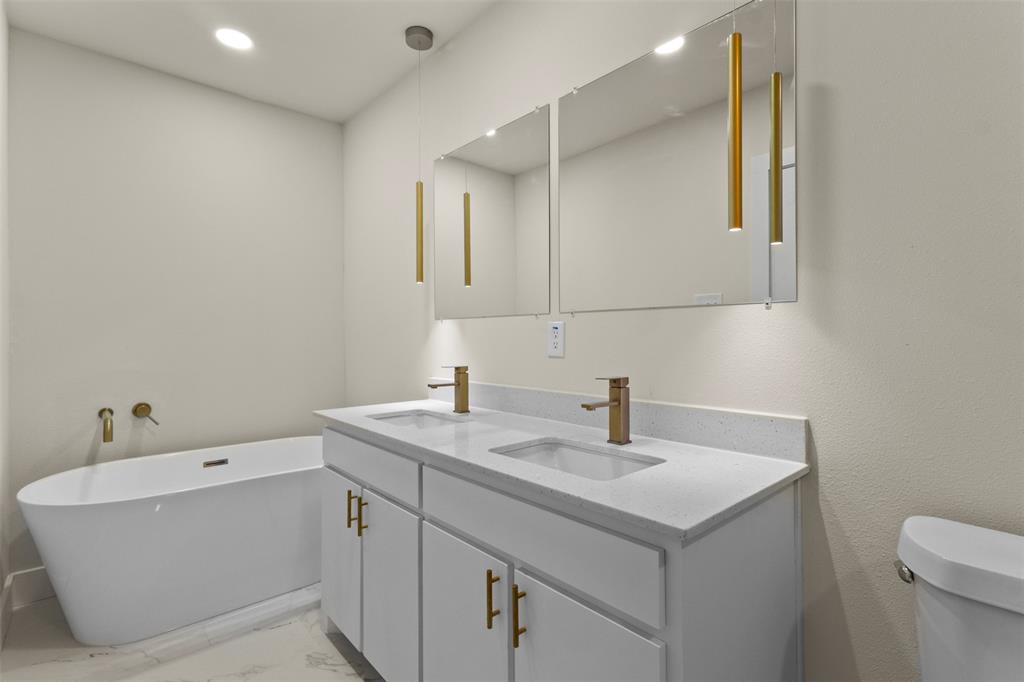 Freestanding soaking tub, dual vanity with undermount sinks, quartz countertops, brushed brass fixtures, and wall-mounted mirrors with integrated lighting