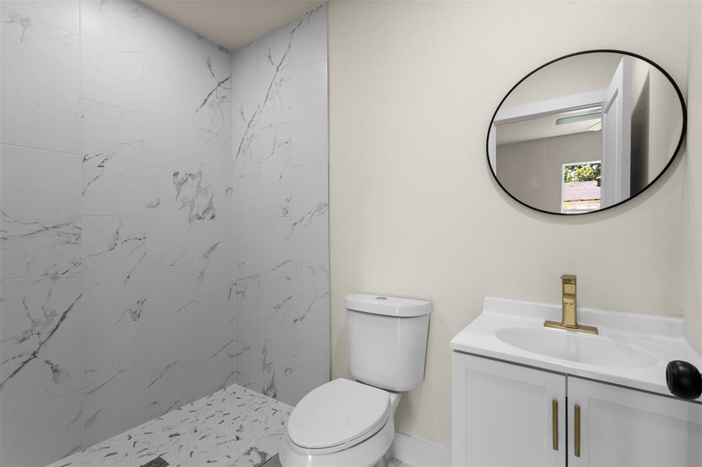 White vanity with gold-tone hardware, a circular mirror, and a walk-in shower with marble-style wall tiles and mosaic floor tiles