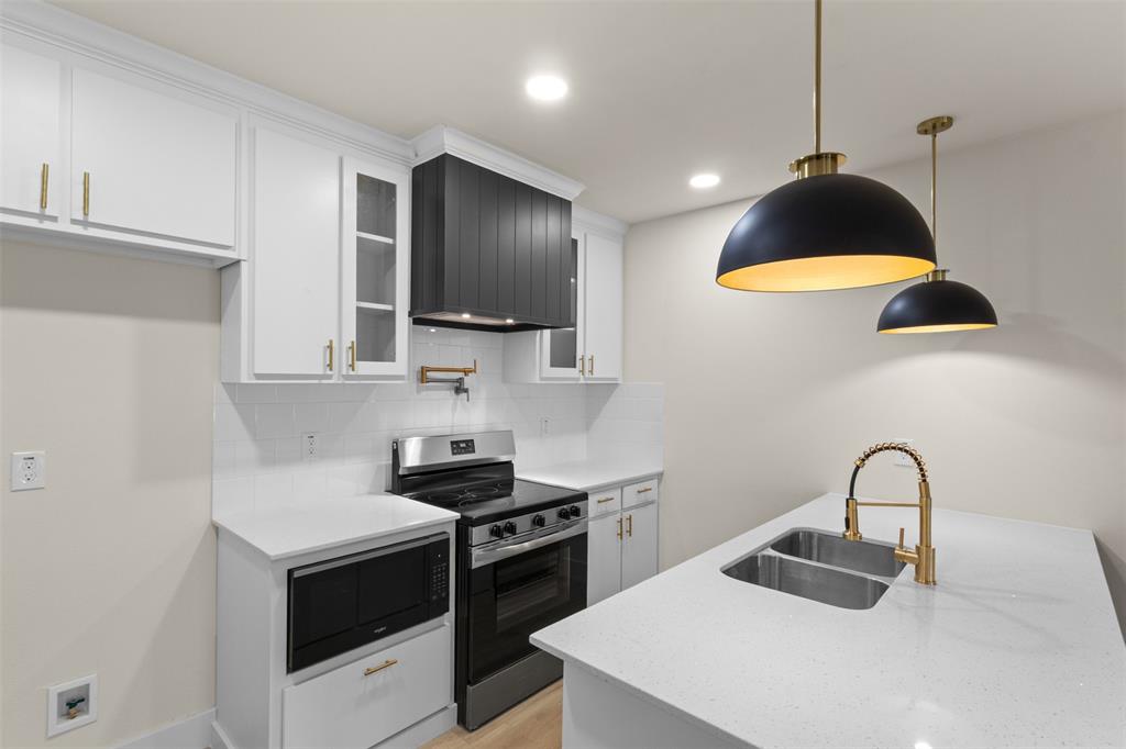 Kitchen featuring white cabinetry, a black range hood, stainless steel appliances, a double basin sink with a gold-finish faucet, and pendant lighting