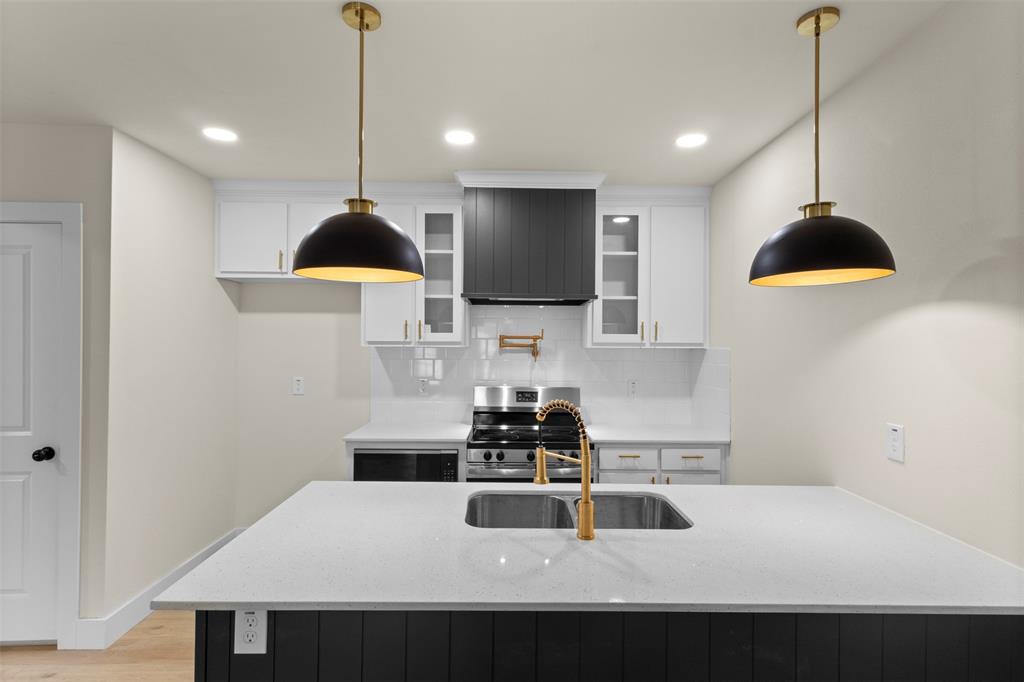 Kitchen island with a light-toned countertop, dual pendant lighting fixtures, a stainless steel sink with a gold-toned faucet, and dark-toned cabinetry