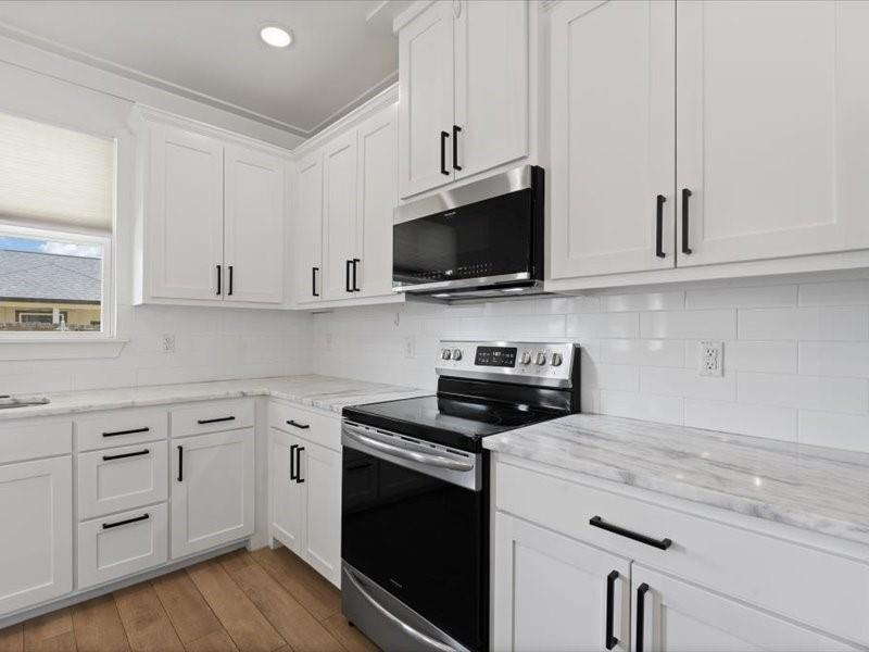 Kitchen featuring white cabinetry with matte black hardware, stone countertops with subtle veining, a white subway tile backsplash, stainless steel appliances, and wood-finish flooring Kitchen featuring white cabinetry with matte black hardware, stone countertops with subtle veining, a white subway tile backsplash, stainless steel appliances, and wood-finish flooring