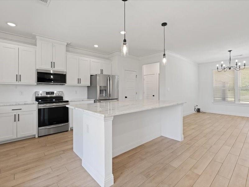 Open concept kitchen featuring an island with a light-toned countertop, white cabinetry with dark hardware, stainless steel appliances, and wood-finish flooring Open concept kitchen featuring an island with a light-toned countertop, white cabinetry with dark hardware, stainless steel appliances, and wood-finish flooring