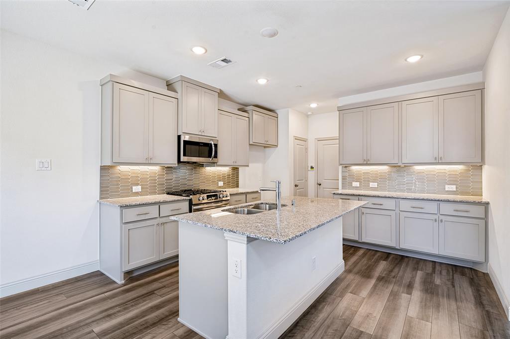 Open-concept kitchen featuring an island with a double sink, light grey cabinetry, stainless steel appliances, granite countertops, and wood-finish flooring
