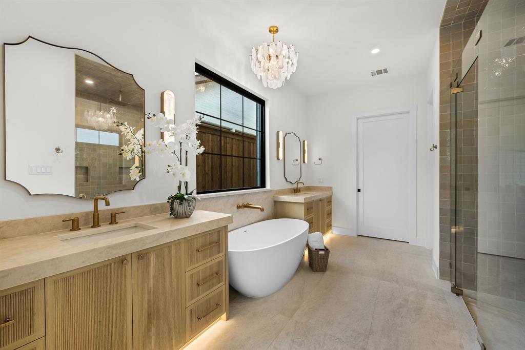 Spacious bath featuring dual wood-finish vanities with light-toned countertops, a freestanding soaking tub, a large walk-in shower with glass enclosure, and a statement chandelier