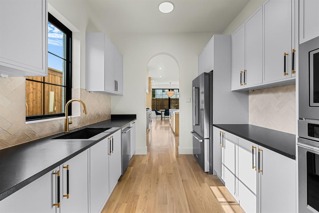 Modern galley kitchen featuring light wood-finish flooring, white cabinetry with contrasting hardware, dark countertops, a herringbone tile backsplash, and a black-framed window