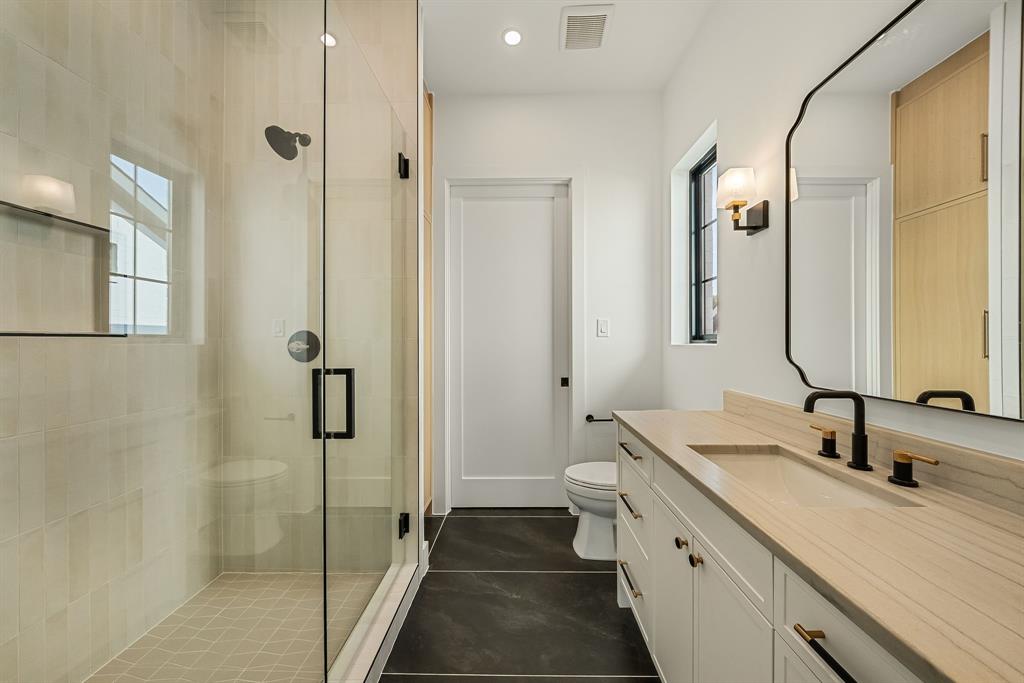 Modern bathroom featuring a frameless glass shower enclosure, a vanity with an integrated sink and light wood-finish countertop, dark tile flooring, a black-framed window, and a decorative wall mirror