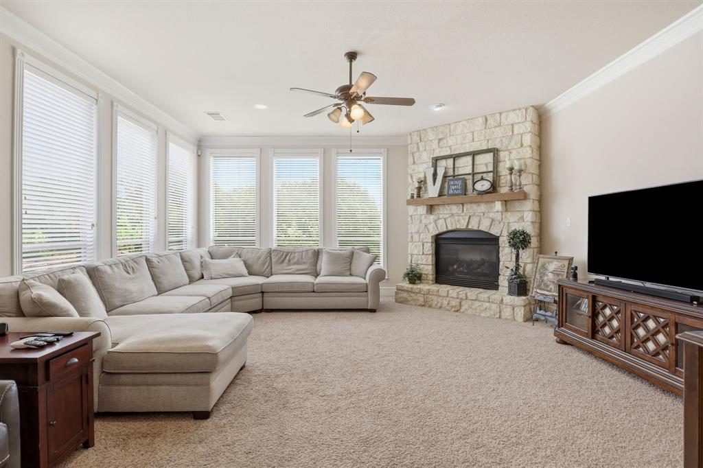 Living area featuring a stone fireplace with a wood mantel, light-colored carpeting, a ceiling fan, crown molding, and multiple windows with horizontal blinds