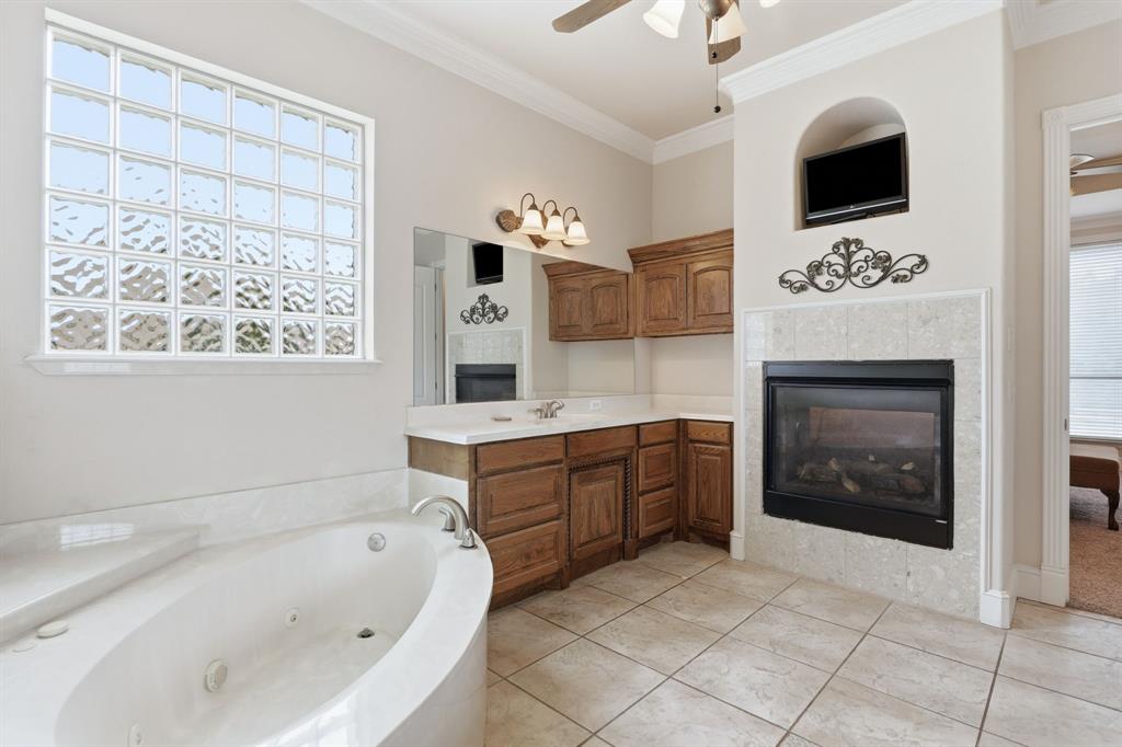 Soaking tub with integrated jets, glass block window, double vanity with wood cabinetry, tiled flooring, and a fireplace with a tiled surround