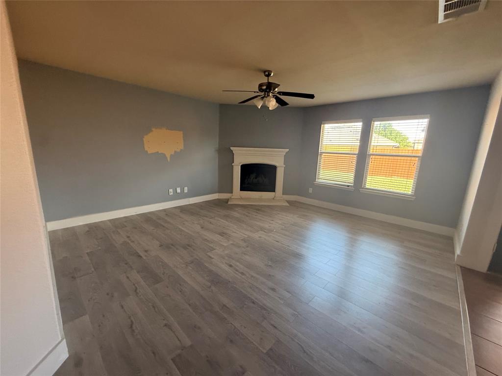 Living area featuring wood-finish flooring, a fireplace with a decorative mantel, multiple windows with blinds, a ceiling fan with integrated lighting, and painted walls Living area featuring wood-finish flooring, a fireplace with a decorative mantel, multiple windows with blinds, a ceiling fan with integrated lighting, and painted walls