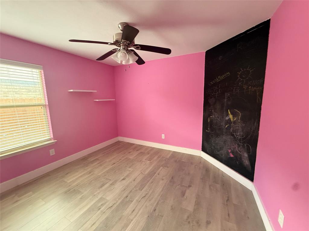 Room featuring wood-finish flooring, a ceiling fan with integrated lighting, a window with horizontal blinds, built-in floating wall shelves, and a chalkboard wall panel