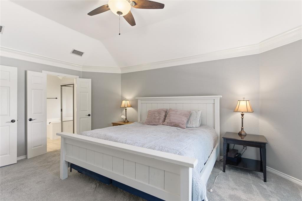 Vaulted ceiling with crown molding, ceiling fan, carpeted flooring, and double doors opening to a bathroom featuring a built-in tub and shower enclosure