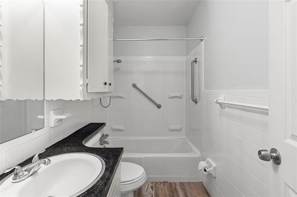 Full bathroom featuring a white tub/shower combination with grab bars, a vanity with a dark countertop and undermount sink, a toilet, and wood-finish flooring