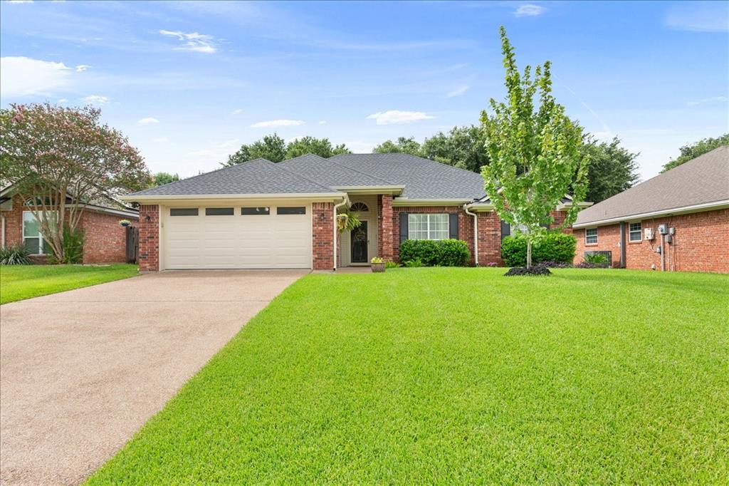 Brick exterior featuring a two-car garage with transom windows, a dark shingle roof, a concrete driveway, and a verdant lawn with mature landscaping