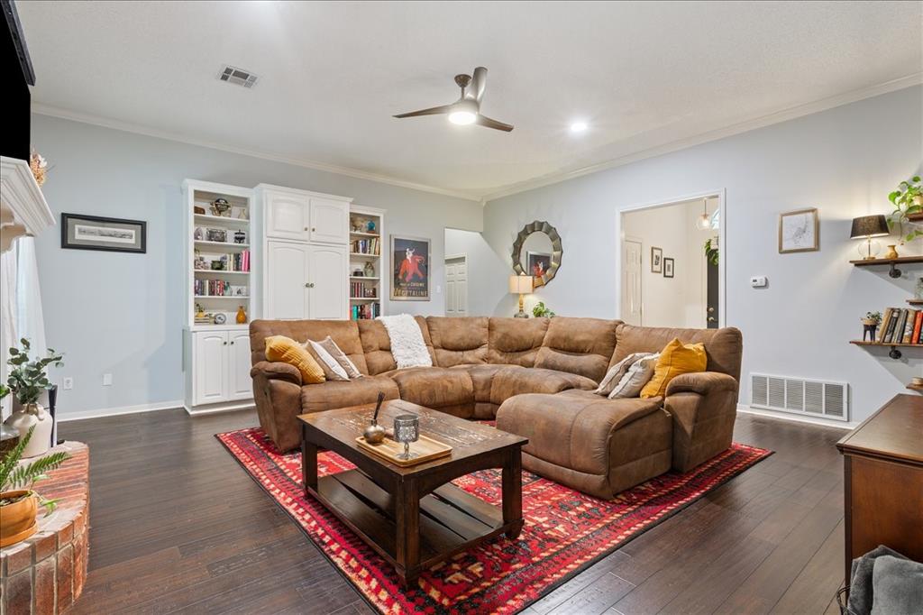 Spacious living area featuring dark wood-finish flooring, built-in white cabinetry, a white crown molding, and recessed lighting Spacious living area featuring dark wood-finish flooring, built-in white cabinetry, a white crown molding, and recessed lighting
