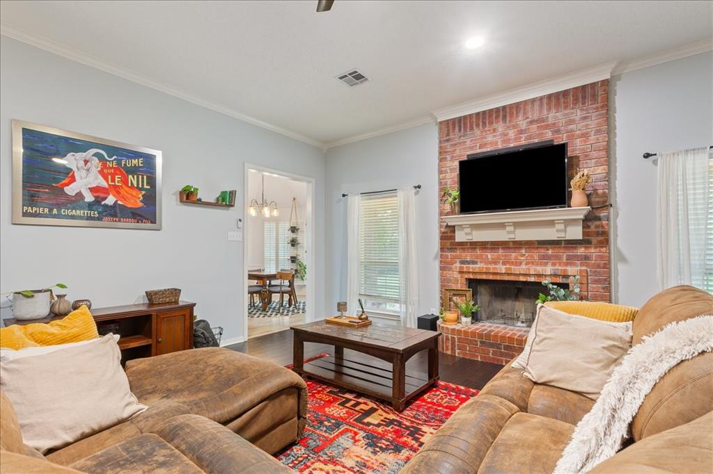 Living area featuring a brick fireplace with a white mantel, dark wood-finish flooring, light-colored walls, crown molding, and a visible doorway to an adjacent room