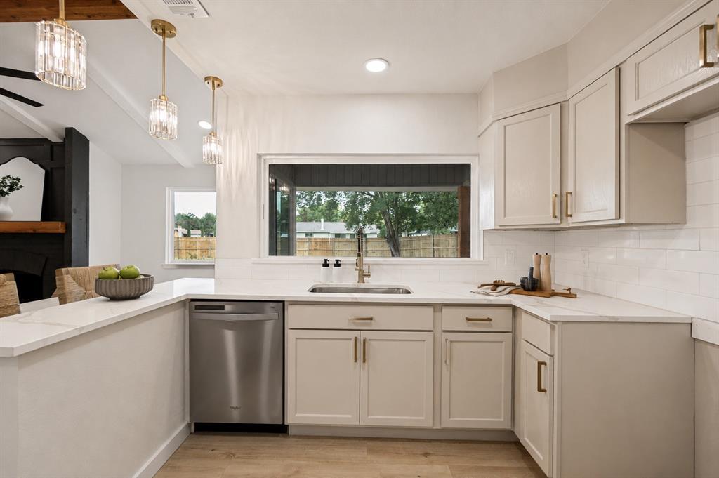 Kitchen featuring light-toned cabinetry with brass hardware, a stainless steel dishwasher, white subway tile backsplash, and light-toned countertops