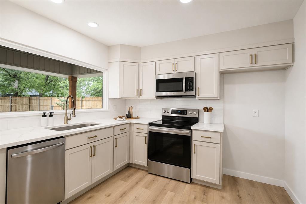 Contemporary kitchen featuring light wood-finish flooring, white shaker-style cabinetry with gold-tone hardware, stainless steel appliances, white subway tile backsplash, and a large window above the sink