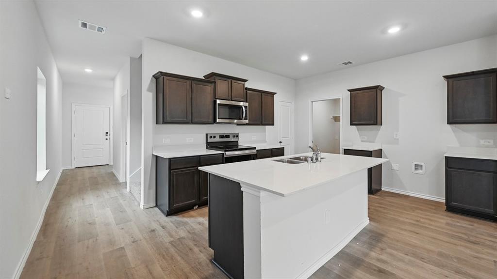 Contemporary kitchen featuring a central island with a double basin sink, dark wood-finish cabinetry, light-toned countertops, and stainless steel appliances