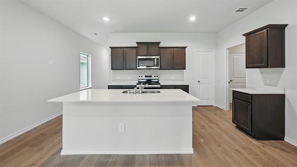 Modern kitchen featuring an expansive island with an integrated sink, dark wood-finish cabinetry, light countertops, stainless steel appliances, and wood-finish flooring