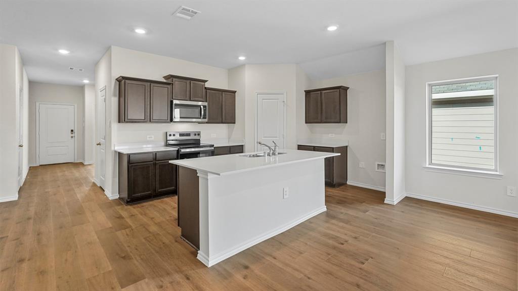 Open-concept kitchen featuring an island with a sink, dark wood-finish cabinetry, stainless steel appliances, white subway tile backsplash, and wood-finish flooring