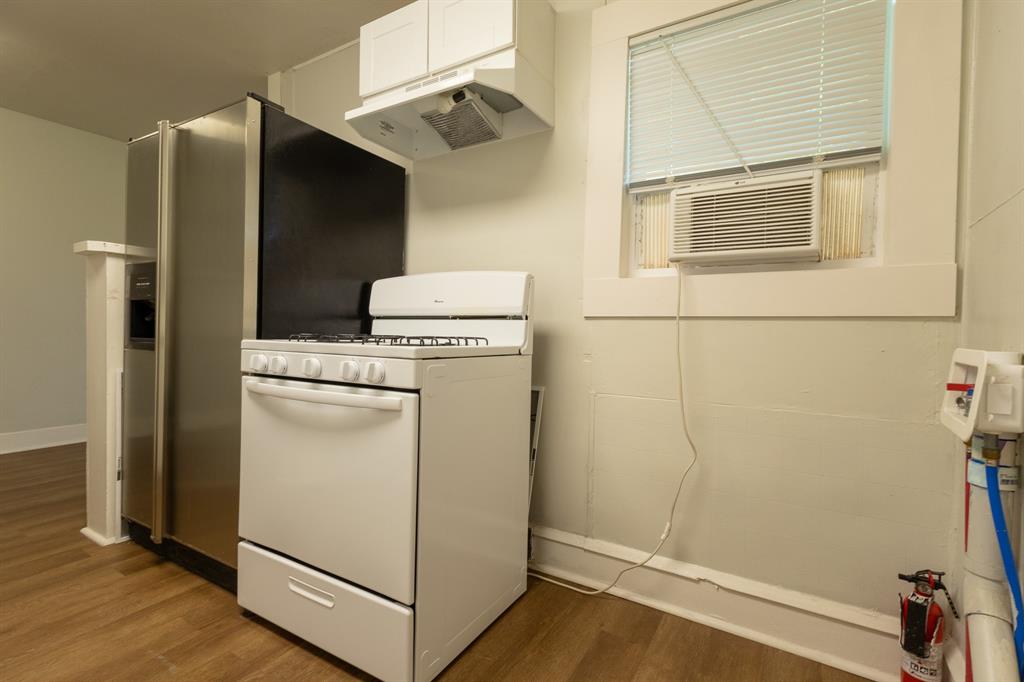 Kitchen area featuring wood-finish flooring, a white gas range with overhead exhaust, stainless steel refrigerator with an ice/water dispenser, and a window with an integrated air conditioning unit Kitchen area featuring wood-finish flooring, a white gas range with overhead exhaust, stainless steel refrigerator with an ice/water dispenser, and a window with an integrated air conditioning unit