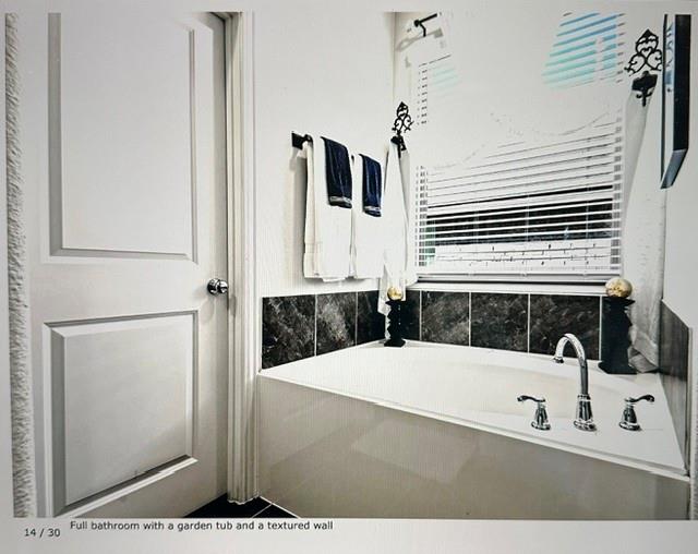 Garden tub with deck-mounted chrome fixtures, surrounded by dark tile accents and a window with horizontal blinds