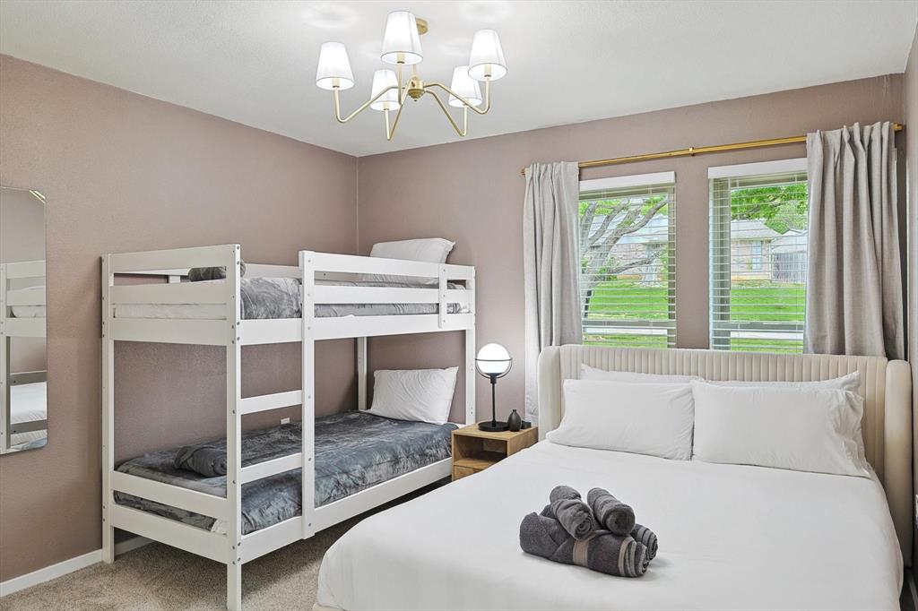 Bedroom featuring two window openings with blinds, a white bunk bed frame, a bed with a cream upholstered headboard, a bedside table, and a contemporary ceiling light fixture