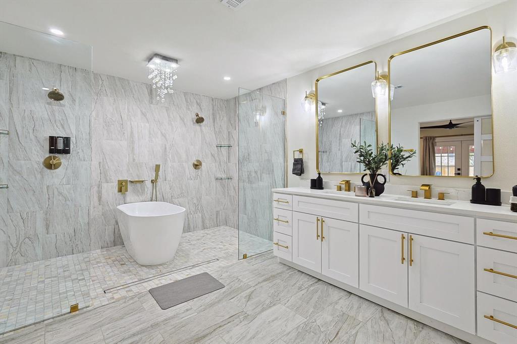 Luxurious bathroom featuring a freestanding soaking tub, a spacious walk-in shower with multiple showerheads, a dual vanity with white cabinetry and gold-toned hardware, and large wall-mounted mirrors with sconce lighting
