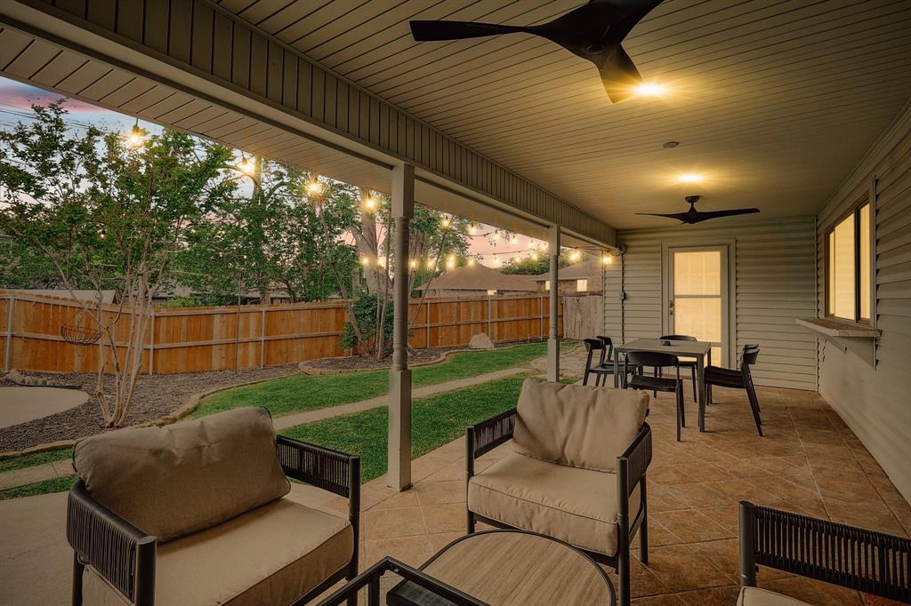 Spacious covered patio featuring ceiling fans, string lighting, tiled flooring, wood fence, and a private backyard with mature trees