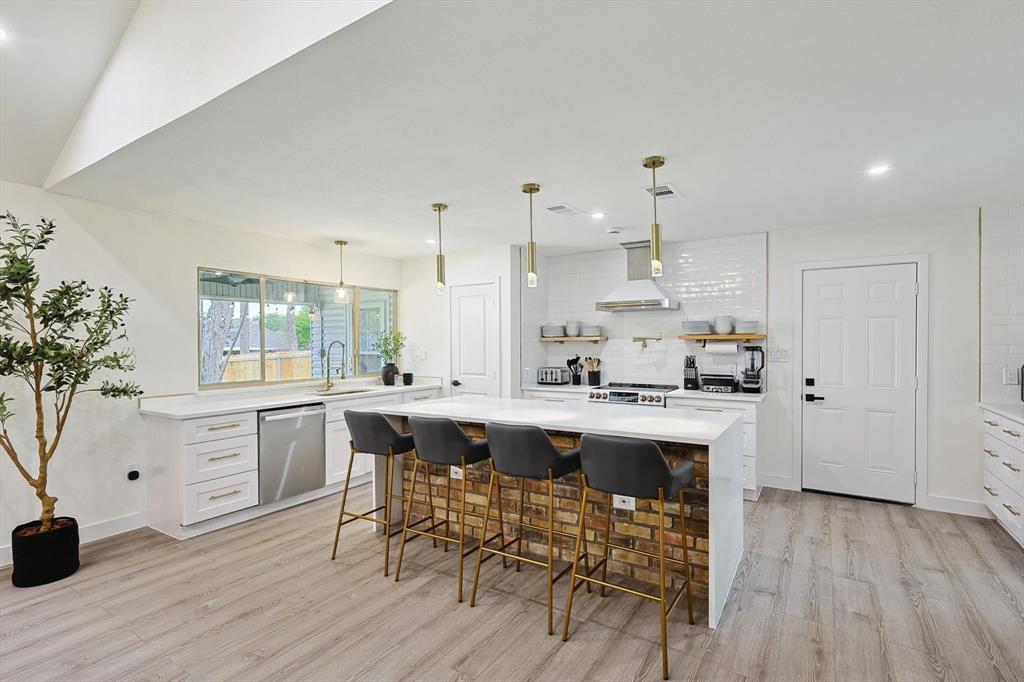 Open-concept kitchen featuring an expansive island with a brick-finish base, integrated stainless steel appliances, white subway tile backsplash, and wood-finish flooring Open-concept kitchen featuring an expansive island with a brick-finish base, integrated stainless steel appliances, white subway tile backsplash, and wood-finish flooring