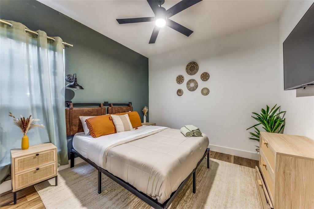 Bedroom featuring an accent wall, window with curtains, wood-finish flooring, ceiling fan with integrated lighting, and a wall-mounted television