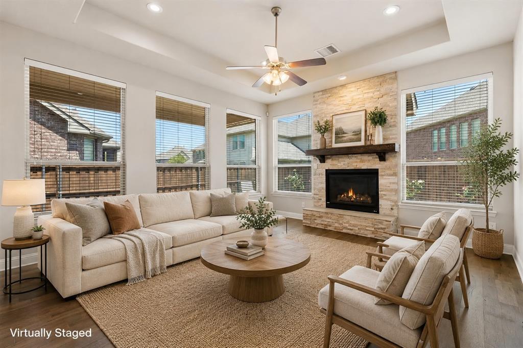 Spacious living area featuring a stone-faced fireplace with a wood mantel, multiple large windows, a ceiling fan, and wood-finish flooring Spacious living area featuring a stone-faced fireplace with a wood mantel, multiple large windows, a ceiling fan, and wood-finish flooring